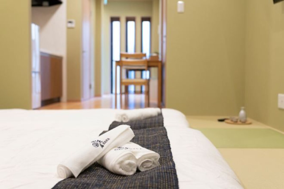 A neatly made bed with rolled towels and a small table with a bottle and a remote control.