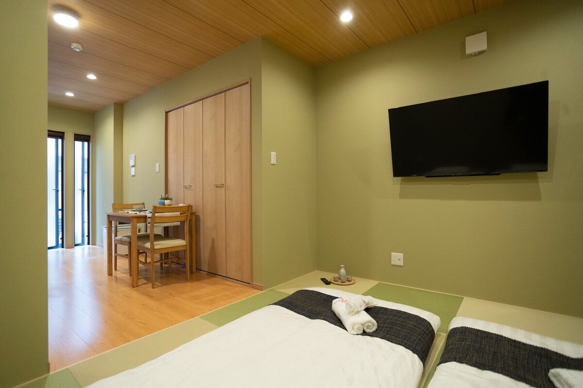 A hotel room with two beds, a TV mounted on the wall, a wooden door, and a dining area with a table and chairs.