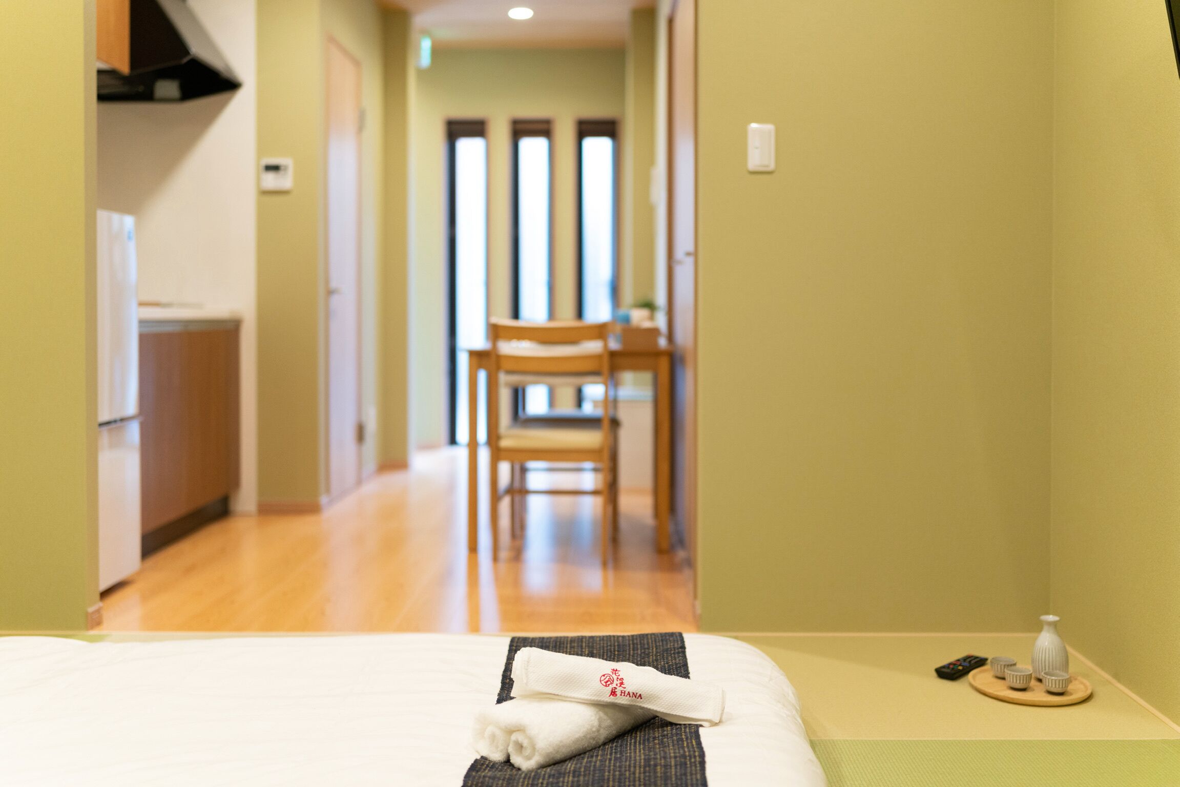 A hotel room with a bed, a small table with a remote control, and a chair in the background.