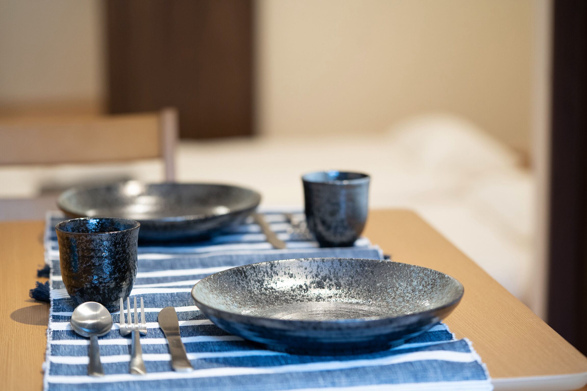 Une table dressée avec une nappe à rayures bleues et blanches, un bol bleu foncé, un bol plus petit, une tasse, une cuillère et une fourchette.