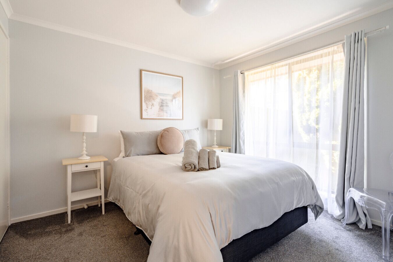 A bedroom with a large bed, two bedside tables with lamps, a framed picture on the wall, and a window with curtains.