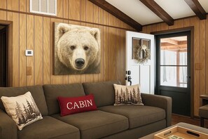Ein gemütliches Blockhaus-Interieur mit einer Holzwand, einem Bären-Motivbild, einer Couch mit Waldkissen und einer Tür mit einem Kranz.