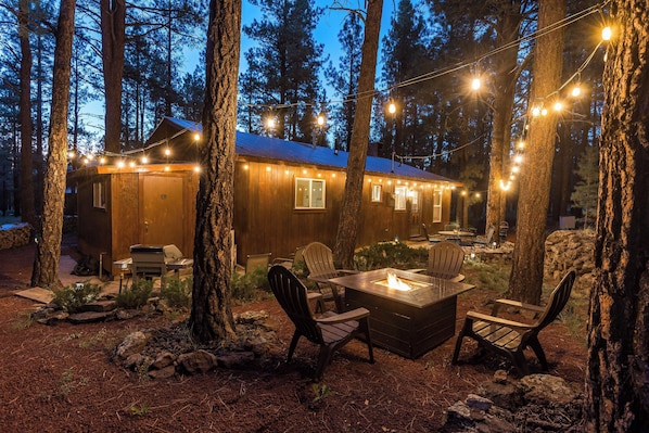 Eine rustikale Holzhütte mit Feuerstelle und Sitzgelegenheit im Freien, umgeben von Bäumen und Lichterketten.