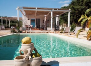 A pool with a lemon in a ceramic jug, a covered outdoor area with lounge chairs, and a building in the background.