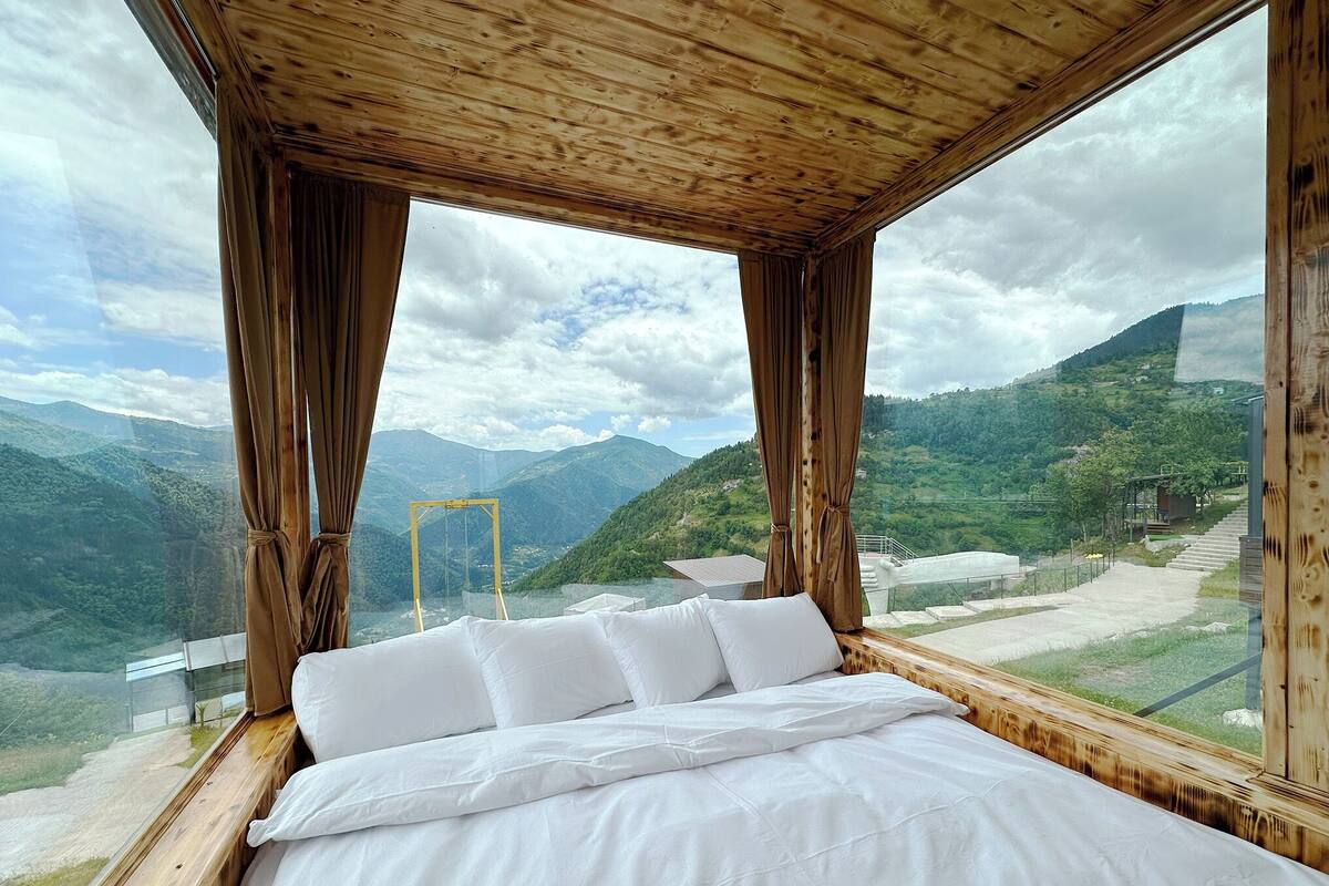Quarto com cama, teto de madeira e janelas amplas com vista para as montanhas.