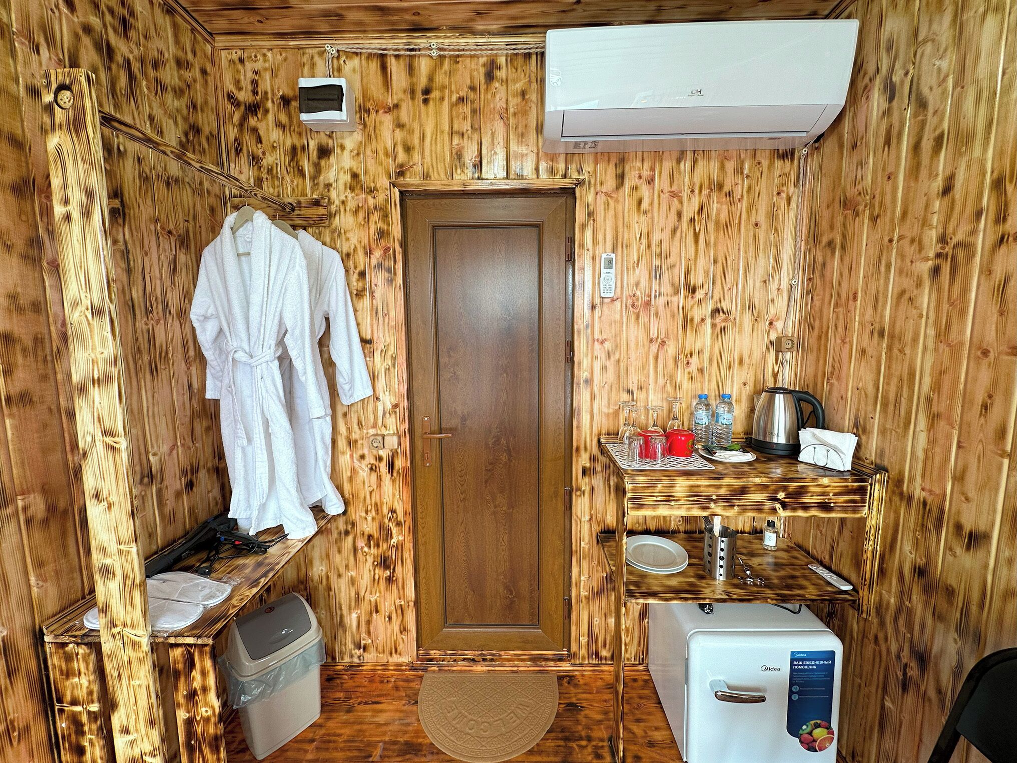 Una habitación de madera con una puerta, una mesita pequeña con vasos y una tetera, y un refrigerador blanco.