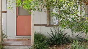 A wooden deck leading to a door, surrounded by lush greenery and a window with a view.