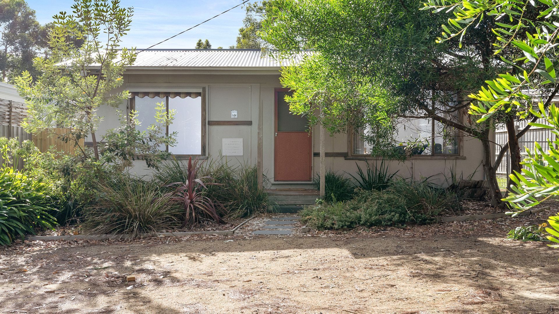 A small house with a front porch, a wooden fence, and a variety of plants and shrubs around it.