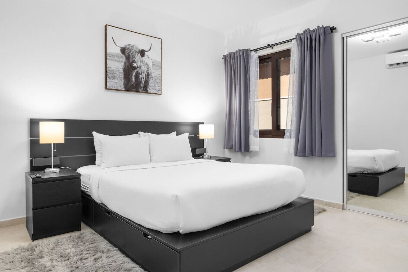 A modern bedroom with a large bed, a headboard, two bedside tables with lamps, a fluffy rug, a window with curtains, and a mirror.