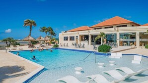 Zona de piscina con sillas de descanso, palmeras y un edificio con tejado de azulejos.