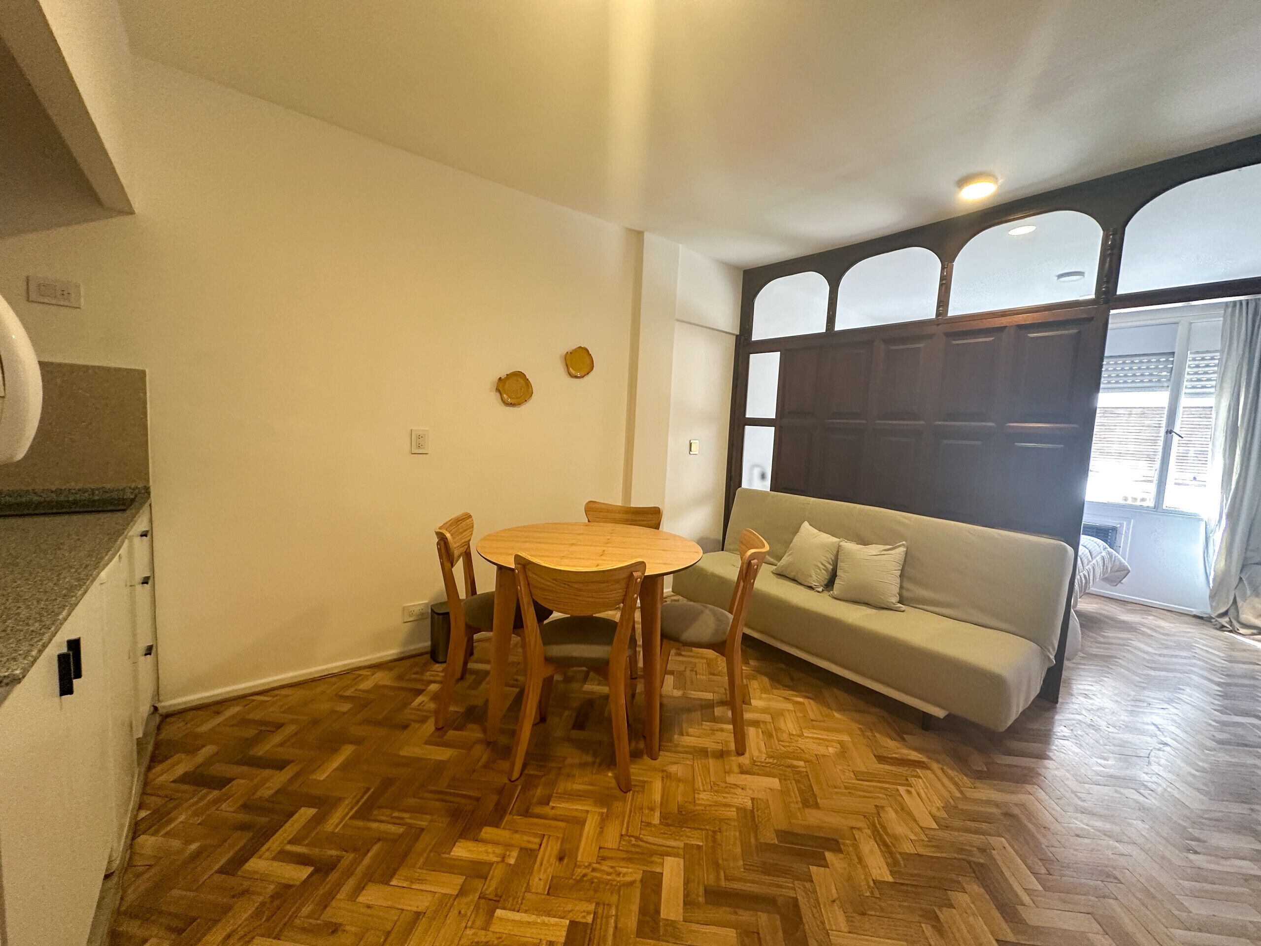 Ein Zimmer mit Holzboden im Fischgrätmuster, einem runden Holztisch mit Stühlen, einem beigen Sofa und einem großen Fenster mit Blick.