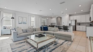 A modern living room with a sectional sofa, a glass coffee table, and a kitchen area with white cabinets and a refrigerator.