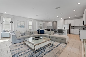 A modern living room with a sectional sofa, a glass coffee table, and a kitchen area with white cabinets and a refrigerator. - 6BR house with pool table near Disney area (Kissimmee)