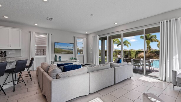 A modern living room with a large sectional sofa, a dining area with a kitchen in the background, and a sliding glass door leading to a pool area.