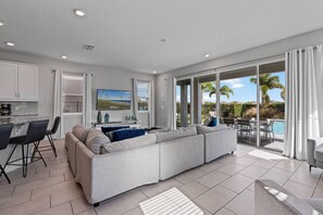Un salon moderne avec un grand canapé d’angle, un coin repas donnant sur une cuisine en arrière-plan, et une porte-fenêtre coulissante qui mène à une zone piscine.
