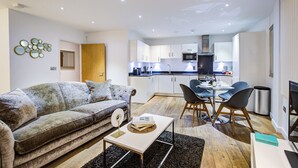 A modern living room with a sofa, coffee table, and dining area with chairs. A kitchen is visible in the background.