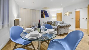 A modern living room with a glass dining table, blue chairs, a sofa, and a painting on the wall.