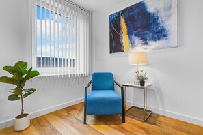 A room with a blue armchair, a potted plant, a table with a lamp, and a large abstract painting on the wall.
