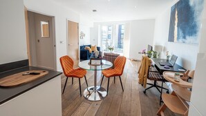 Una cocina moderna con una mesa de comedor de vidrio y sillas color naranja, una computadora portátil sobre un escritorio, y vistas de la ciudad a través de amplios ventanales.