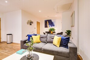 Ein Wohnzimmer mit grauem Sofa, gelben und blauen Kissen, einem weißen Couchtisch und einem Bild an der Wand.