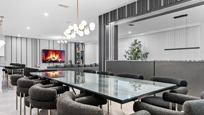A modern conference room with a glass table, grey upholstered chairs, a large TV screen displaying a fireplace, and a potted plant.