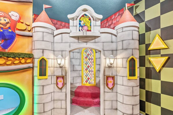 Un espace de jeux sur le thème des châteaux, avec une fresque représentant un personnage de Mario, un vitrail et une entrée recouverte d’un tapis rouge.