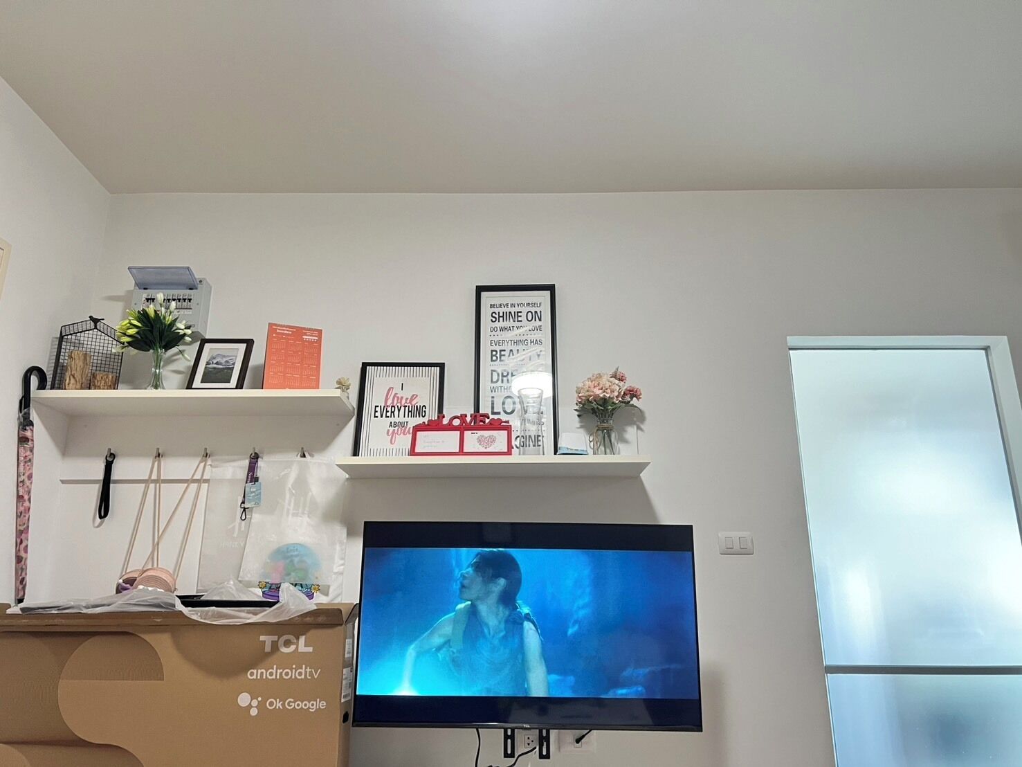 A living room with a flat-screen TV on a stand, a white shelf with decorative items, and a white door.