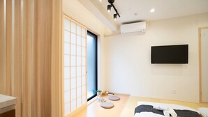 A minimalist hotel room with tatami flooring, a sliding shoji door, a flat-screen TV, and a bed with neatly folded towels. - HANASTAY YAMA (Osaka)
