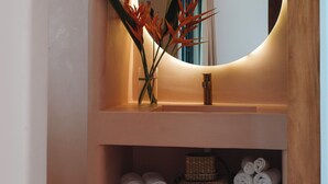 Un tocador de baño con un espejo redondo, un florero con flores rojas y repisas con toallas enrolladas y cestas.