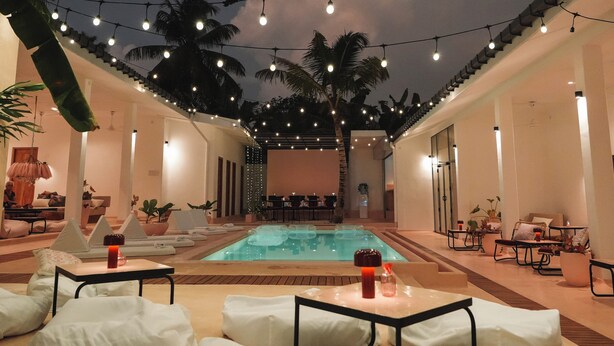 Un área de piscina con sillones de descanso y mesas, luces colgantes y palmeras.