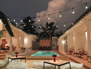 Un área de piscina con sillones de descanso y mesas, luces colgantes y palmeras.