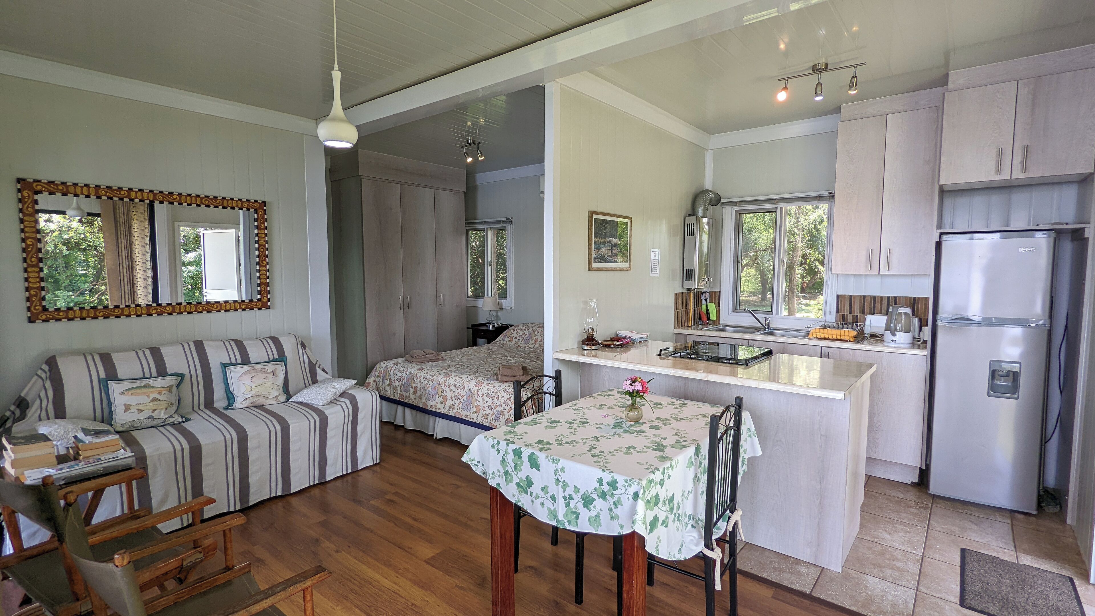 A cozy, well-lit room with a sofa, a small dining table, and a kitchen area with a refrigerator and cabinets.