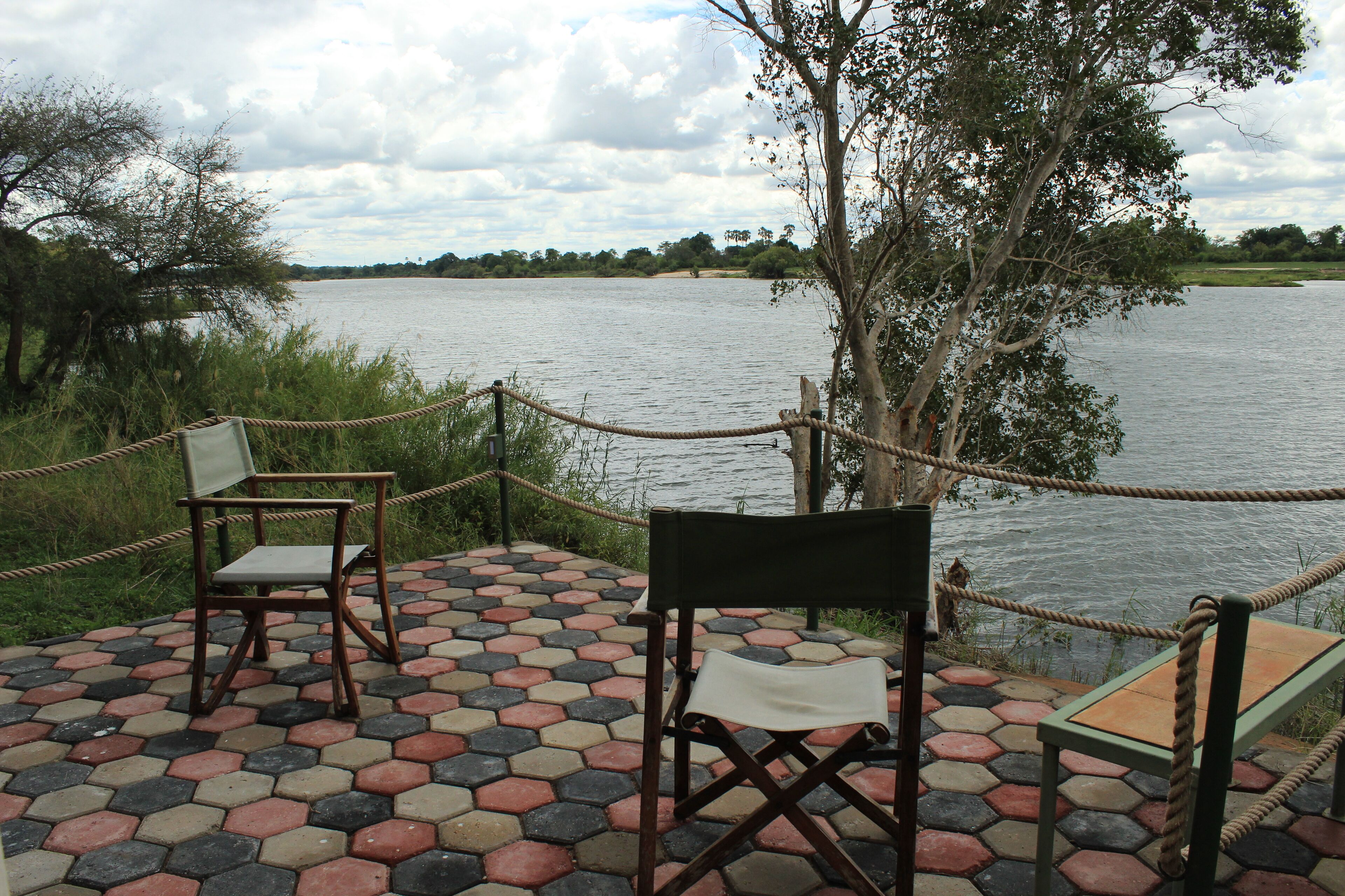 A riverside seating area with hexagonal tiles, two wooden chairs, and a rope barrier.