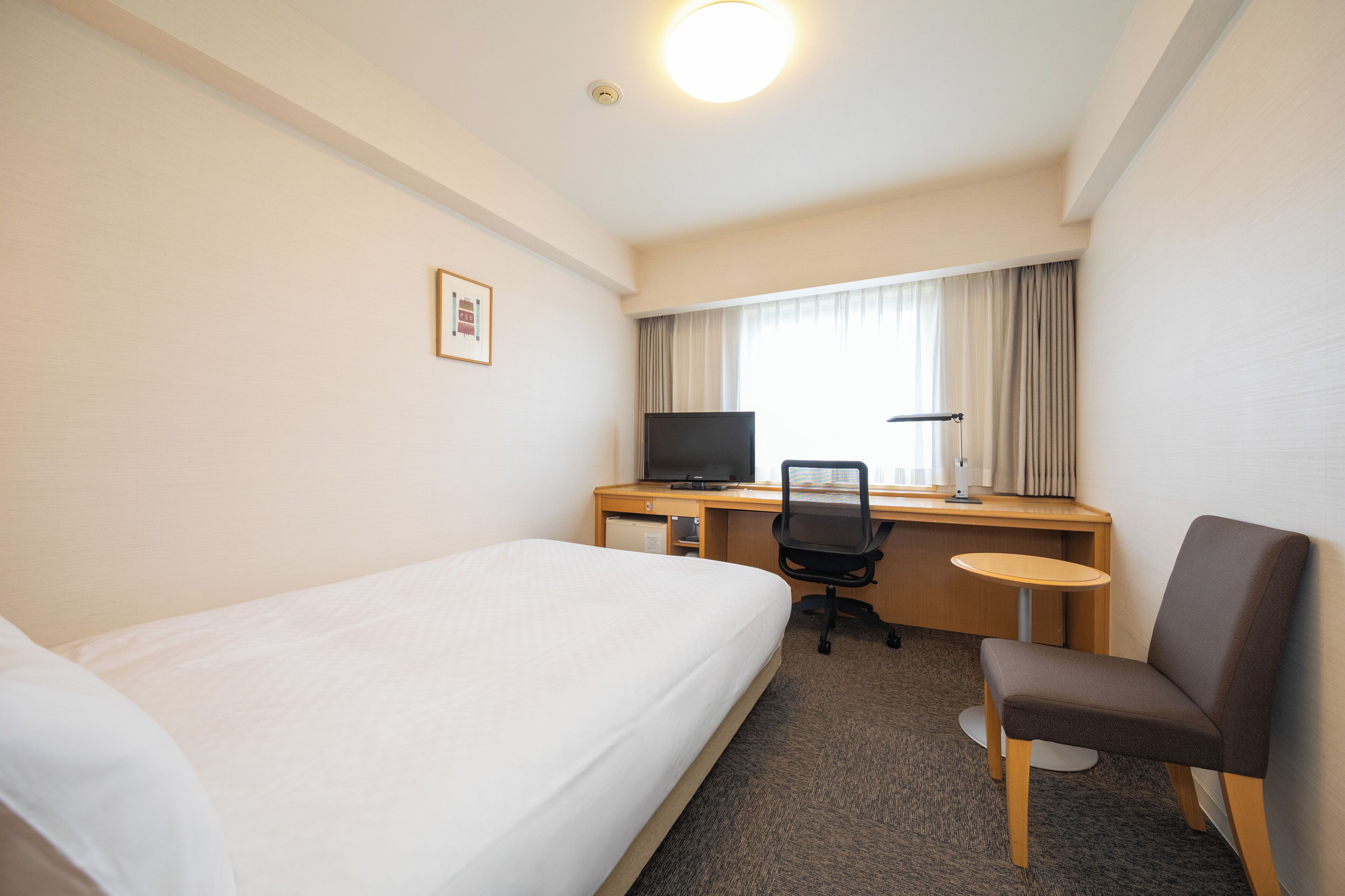 Habitación de hotel con una cama individual, un escritorio con computadora y silla, y una mesita pequeña.