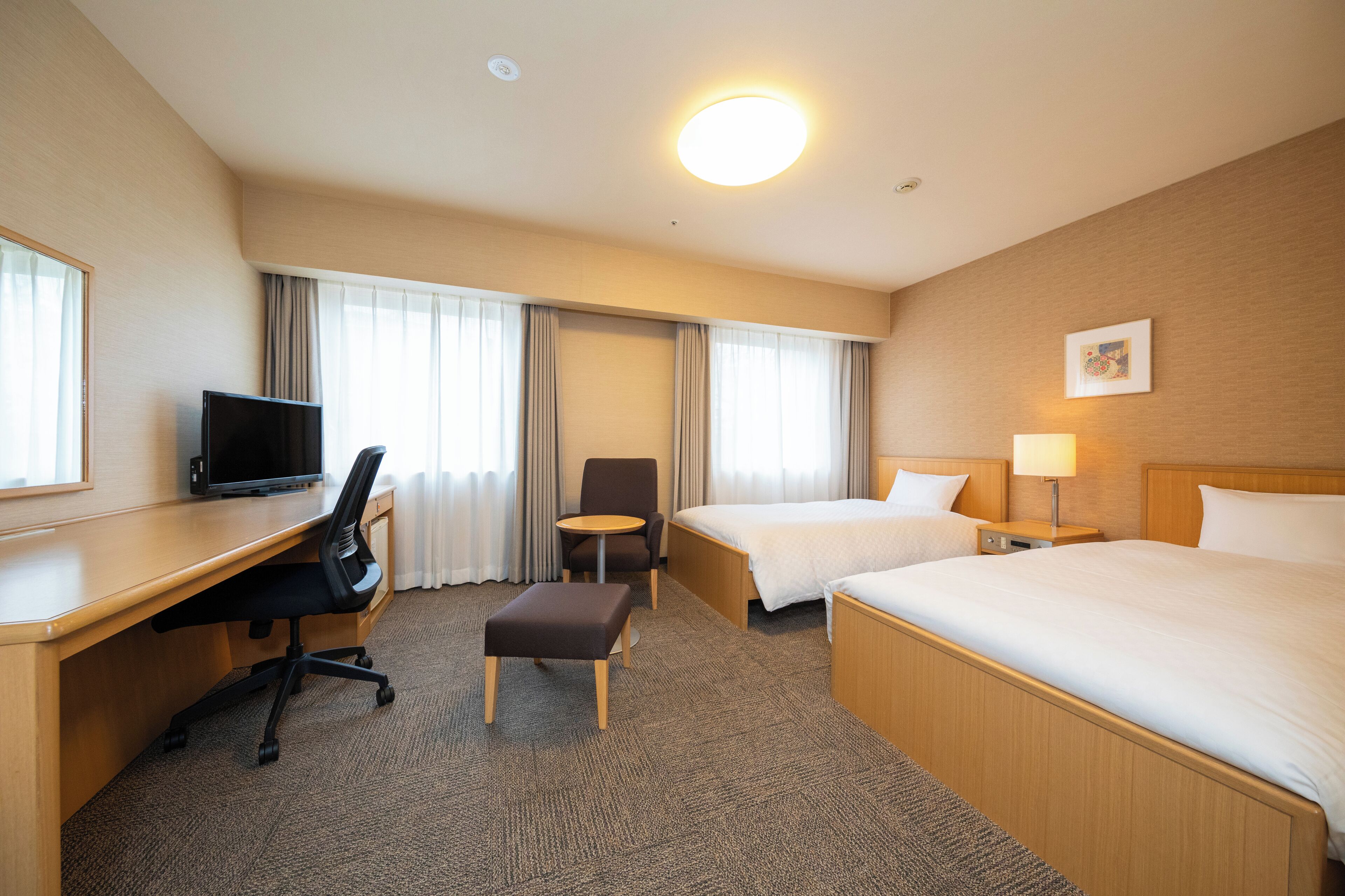 A hotel room with a desk, two beds, a chair, a small table, and a lamp.