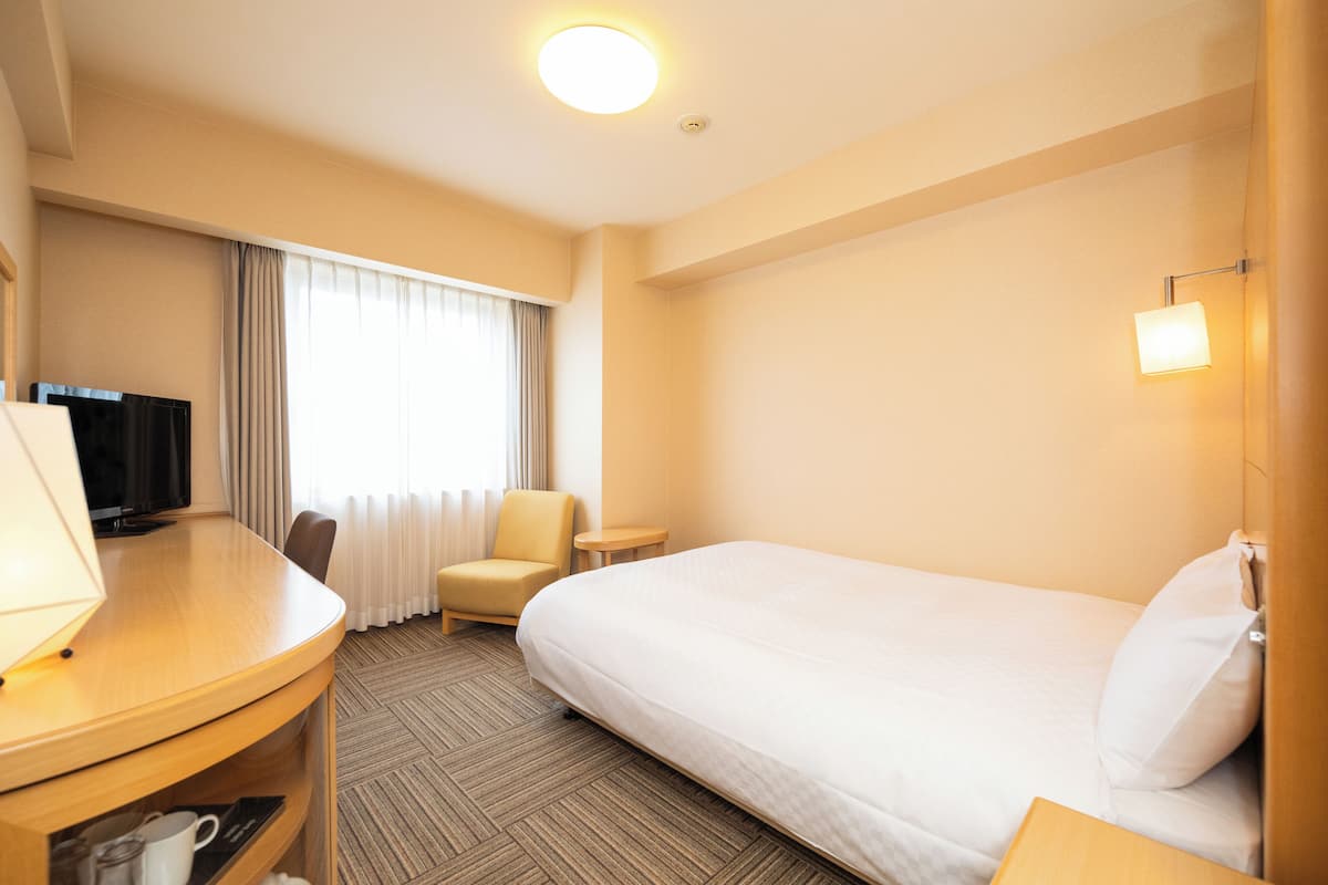 Habitación de hotel con cama, escritorio con televisor, una silla, una mesita y ventana con cortinas.