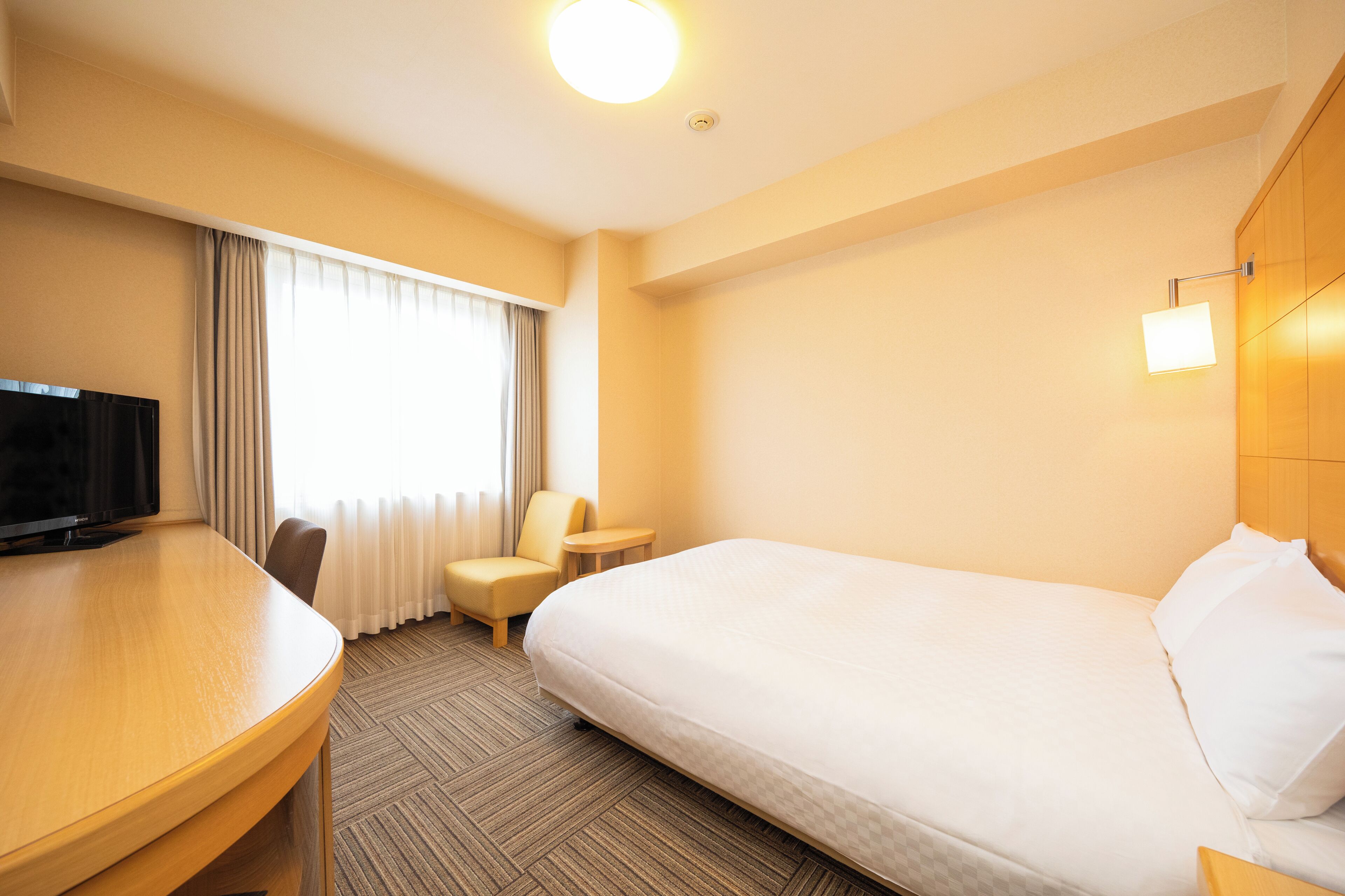 A hotel room with a bed, a desk with a television, a chair, and a window with curtains.