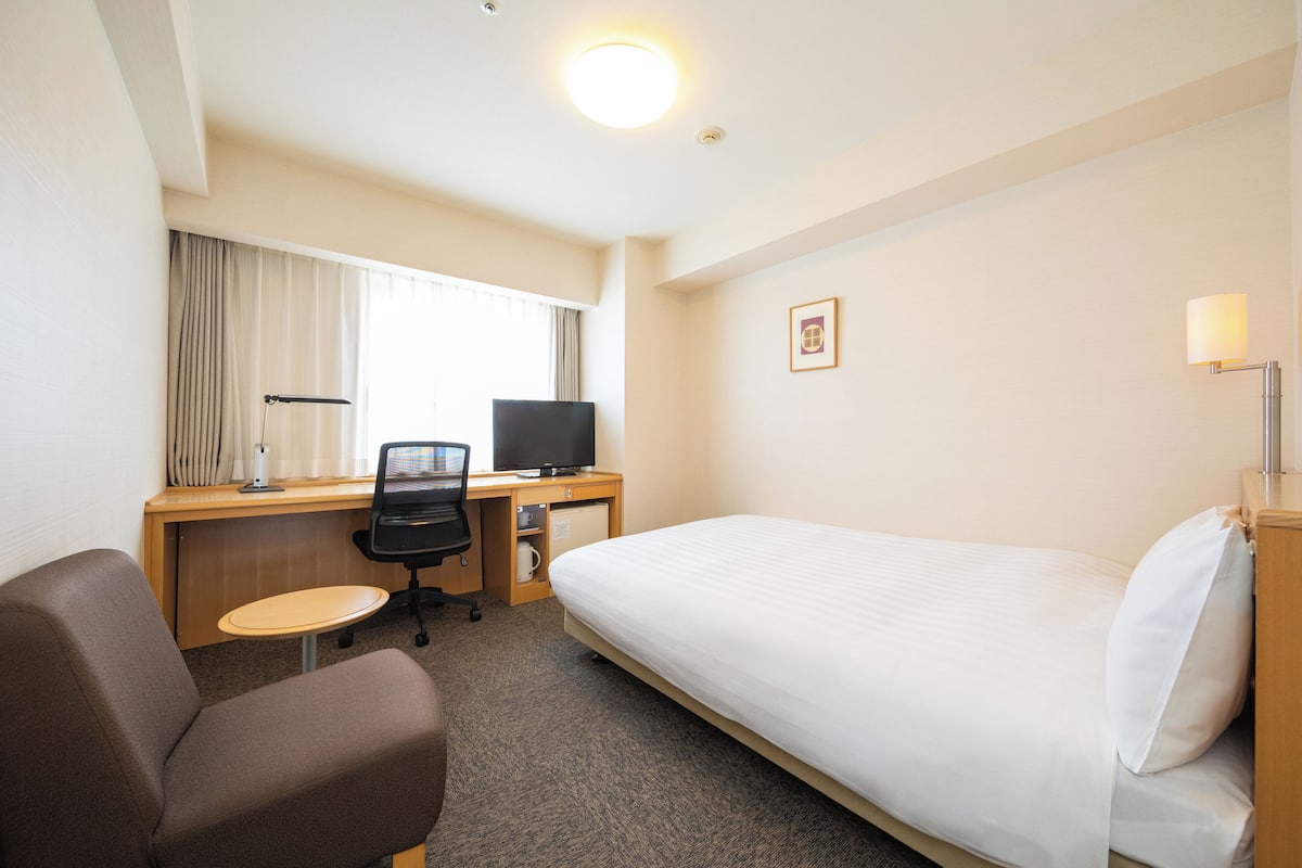 Habitación de hotel con una cama individual, un escritorio con ordenador, una silla y una mesita.