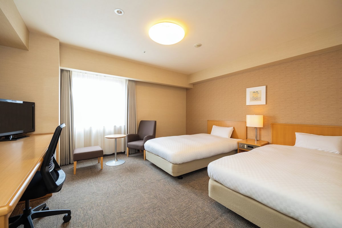 Habitación de hotel con dos camas, un escritorio con silla, un televisor, una mesita redonda y un ventanal con cortinas.