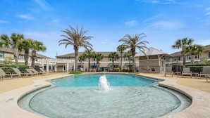 Une piscine avec une fontaine, entourée de palmiers et de chaises longues, avec un bâtiment en arrière-plan.