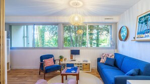 A modern living room with a blue sofa, a pink couch, and a wooden coffee table. There is a TV on a white stand and a large window with a view of trees.