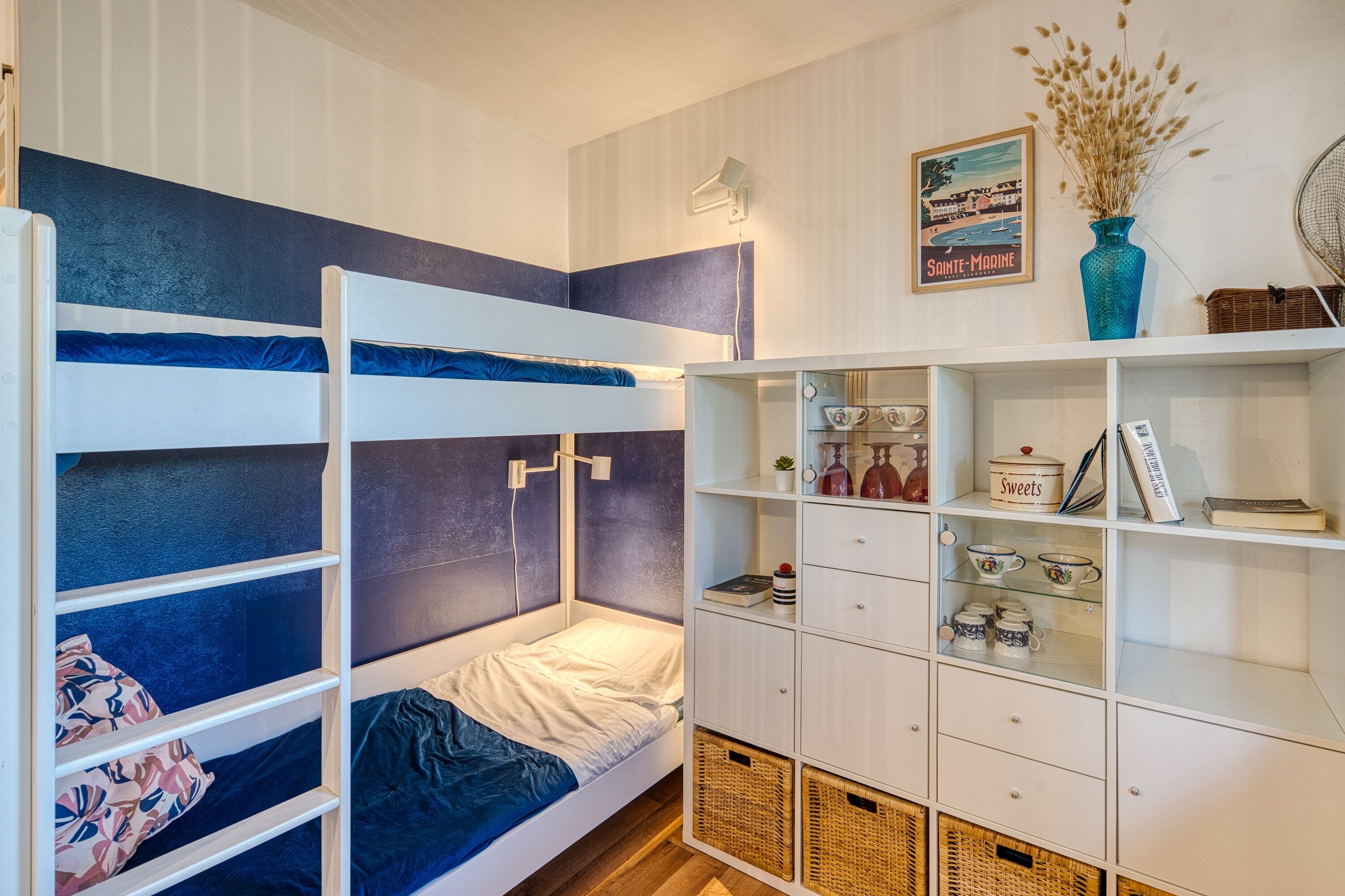 Habitació amb literes, amb una combinació de colors blanc i blau, un petit moble amb prestatges i una fotografia emmarcada a la paret.