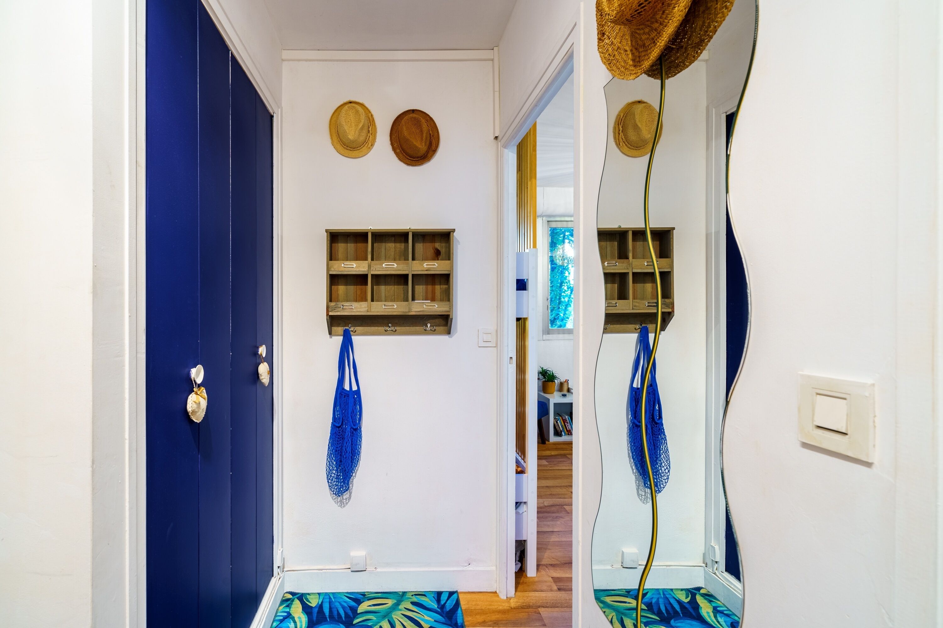 A hallway with a blue door, a wooden shelf with hats, and a mirror reflecting the hallway.