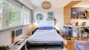 A modern bedroom with a large window, a bed, a desk, and a kitchenette.
