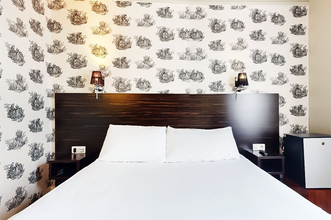 Habitación de hotel con una cama grande, cabecera de madera oscura, dos mesitas de noche con lámparas y un papel tapiz estampado.