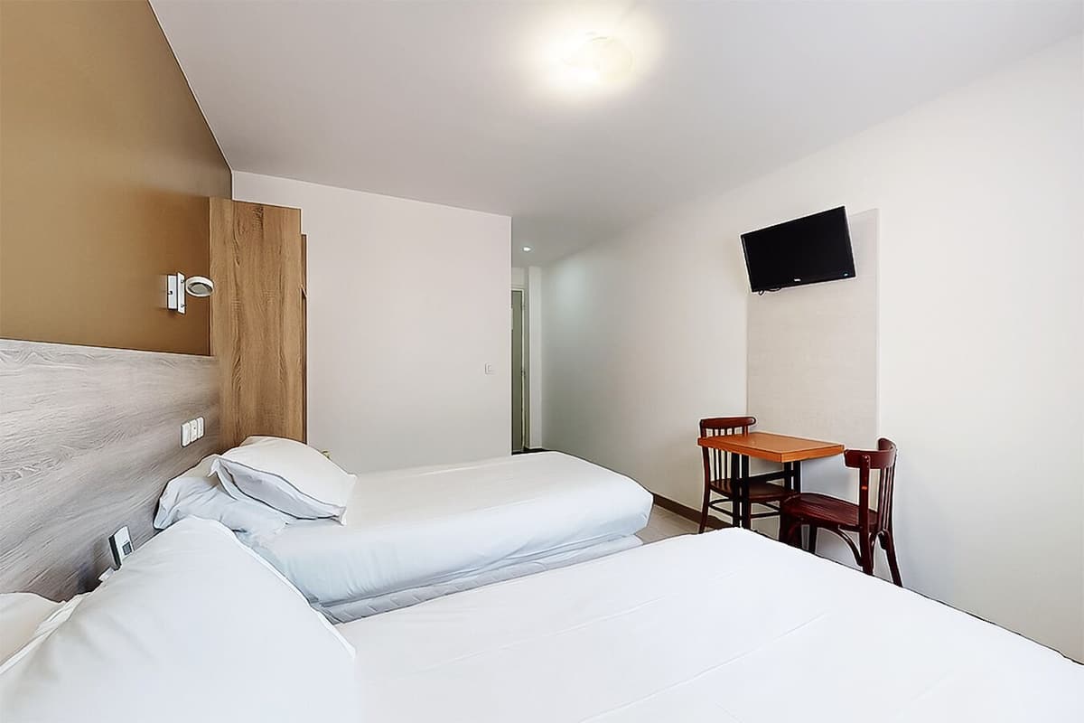 A hotel room with two beds, a wooden wardrobe, a small table with a chair, and a flat-screen TV mounted on the wall.