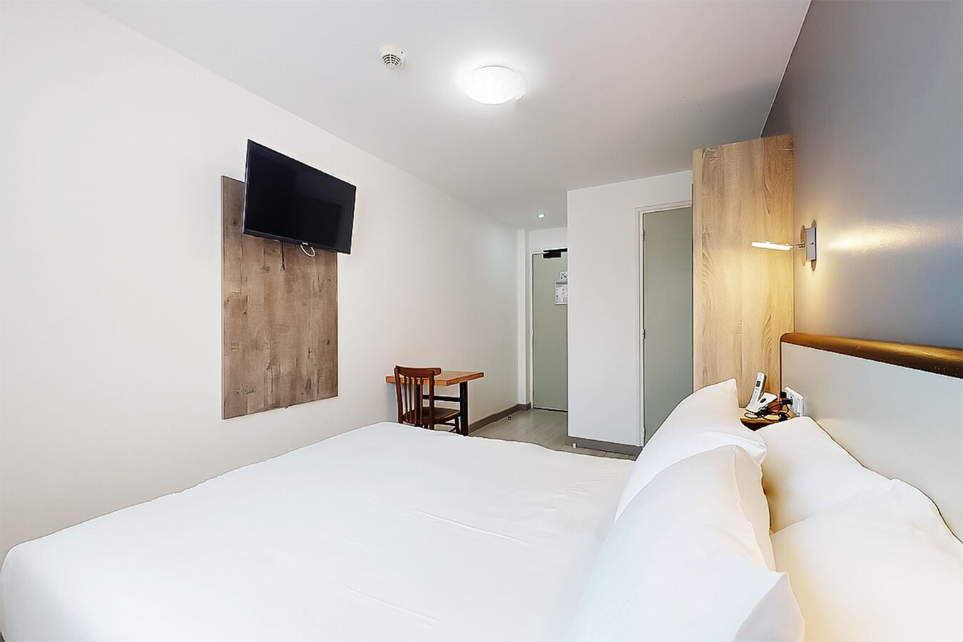 A hotel room with a large bed, a small table, a chair, a TV mounted on the wall, and a wooden door.