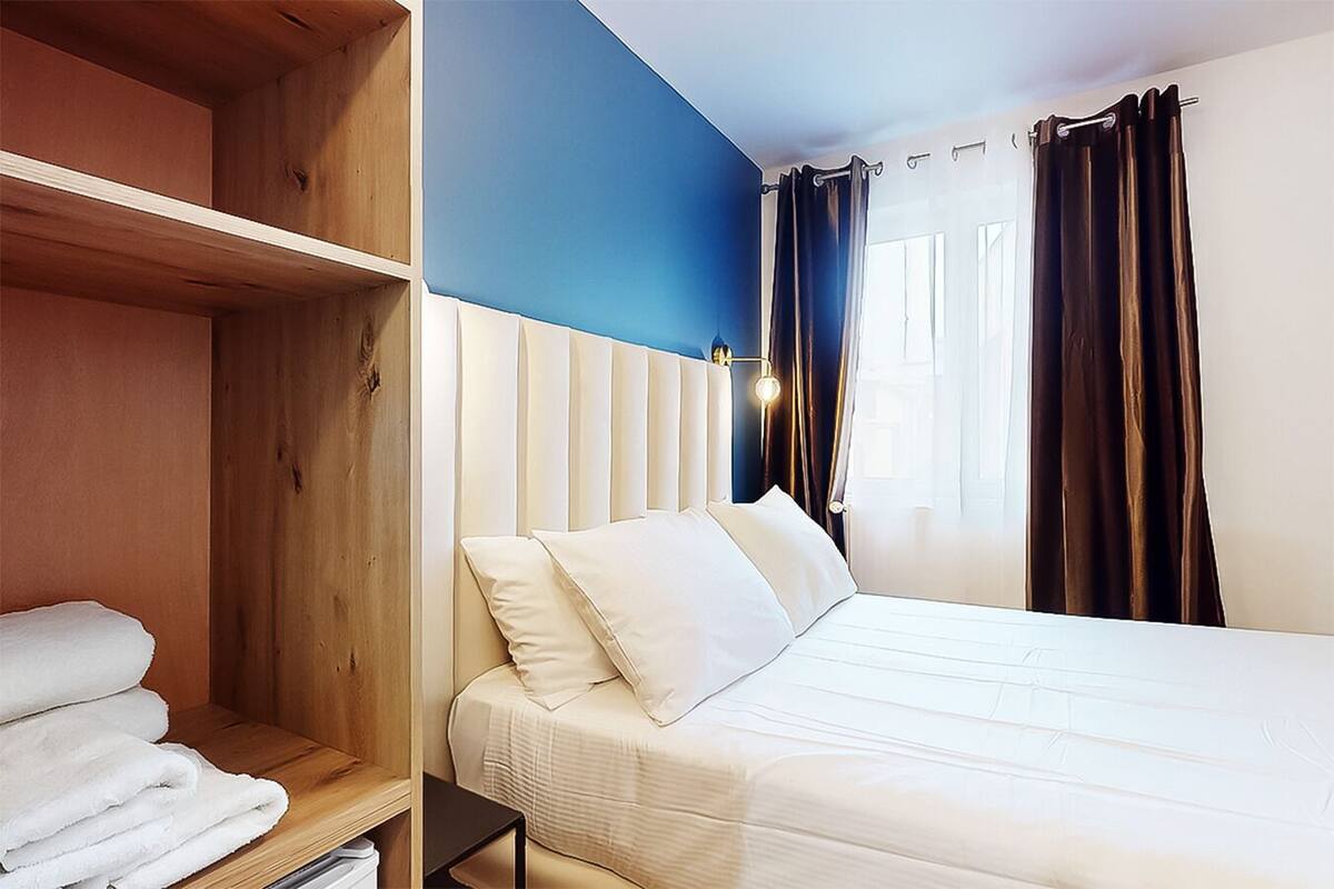 A hotel room with a bed, a wooden shelf with towels, a blue accent wall, and dark curtains.