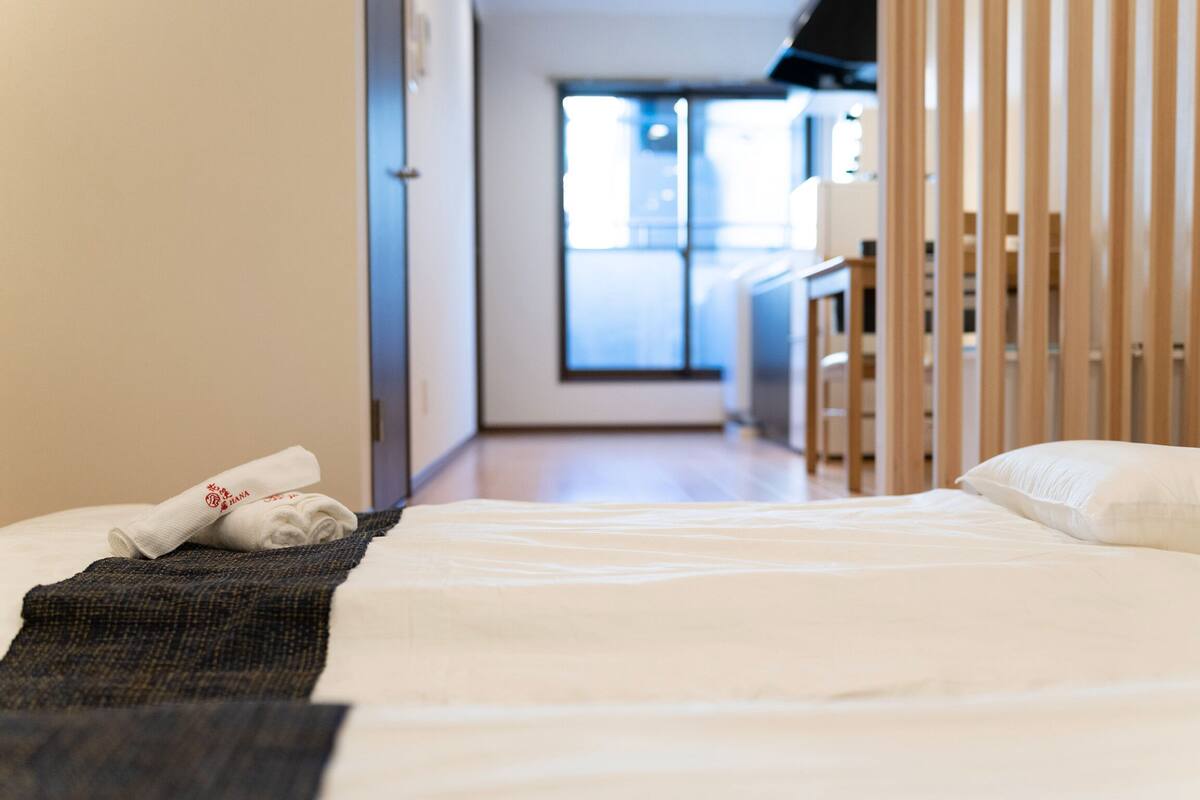 A hotel room with a neatly made bed, a small bedside table, and a view of the hallway and another room through the door.