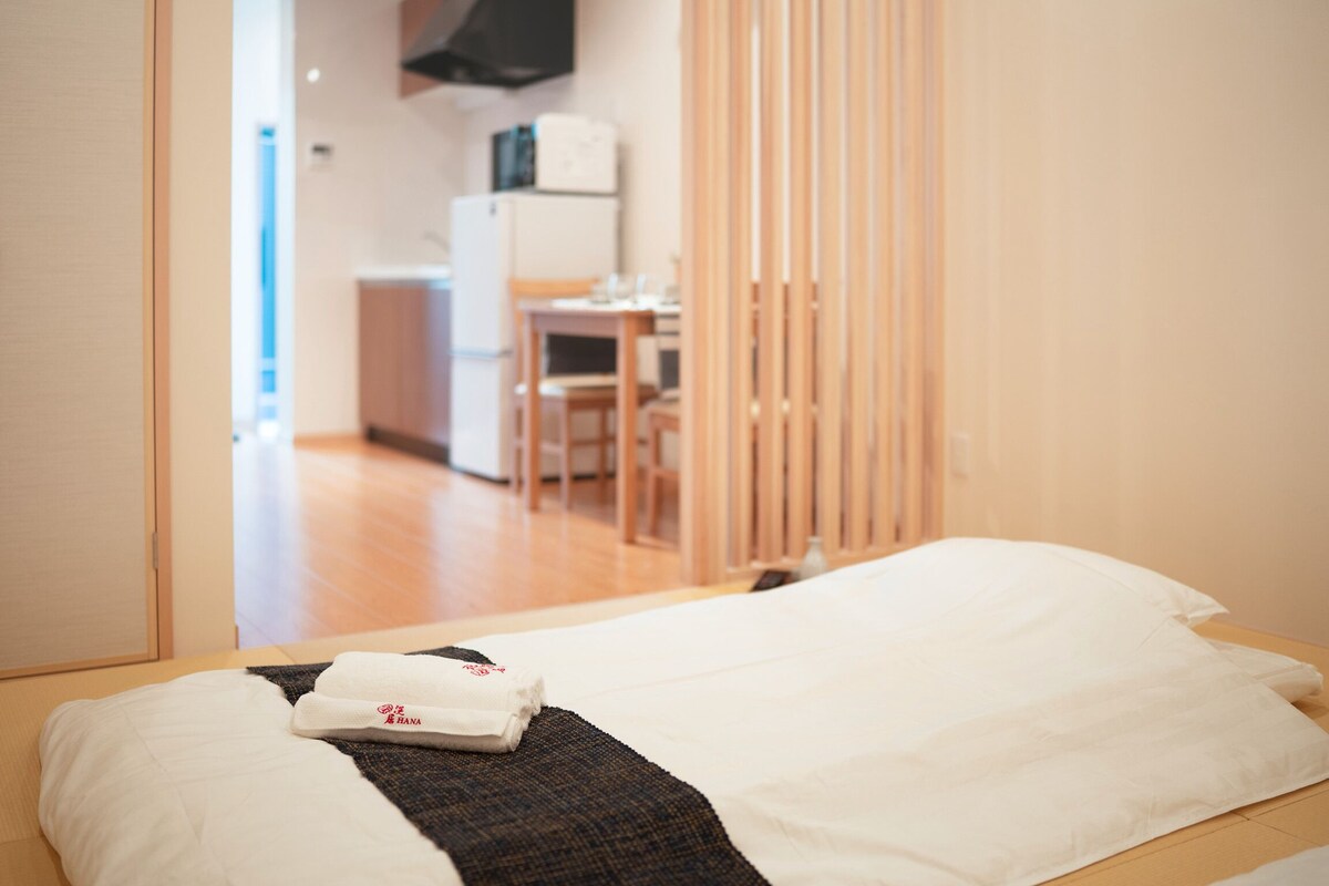 A hotel room with a bed, a neatly made bed with a white pillow and a folded towel, a wooden floor, and a view of a kitchen area with a table and chairs.